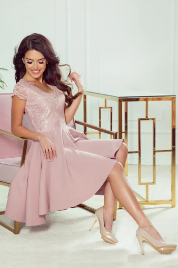 Modelka prezentuje elegancką sukienkę z koronkową górą, która idealnie podkreśla kobiecość i styl. Sukienka w delikatnym odcieniu różu doskonale komponuje się z klasycznymi szpilkami, tworząc harmonijną całość na każdą okazję.