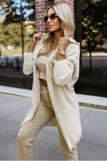 The woman in the photo is wearing an elegant ivory knit cardigan, styled with a matching cream tank top for a cohesive look. The knit cardigan drapes gracefully over her shoulders, complementing her light-colored trousers and adding an element of sophistication to the ensemble.