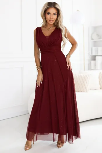 The model showcases an elegant sleeveless burgundy evening gown with a flowing skirt. This burgundy gown features a ruched bodice, adding a touch of sophistication to the overall design.