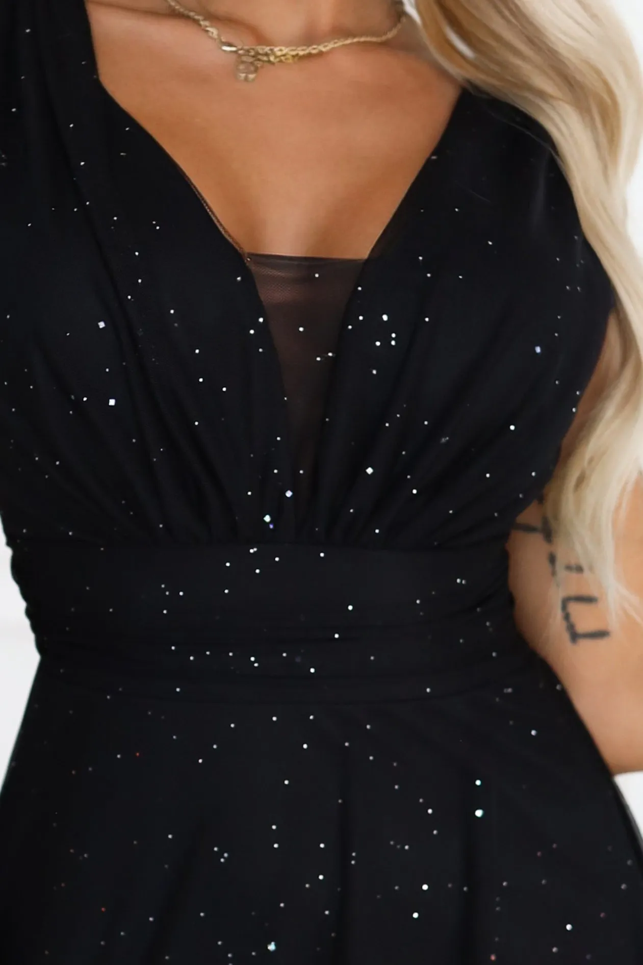 The model showcases a black sparkling evening dress adorned with delicate shimmering sequins. This glamorous gown features a deep V-neckline and subtle draping for an elegant appeal.